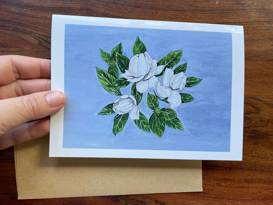 Watercolor painting of white gardenia and  green leaves with blue background on a greeting card that is blank inside.