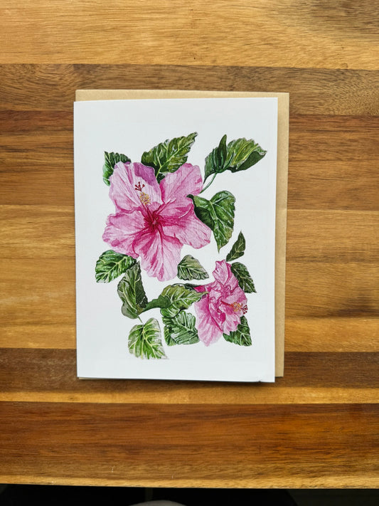 Watercolor pink hibiscus flowers with some green foliage leaves on a greeting card that is blank inside.